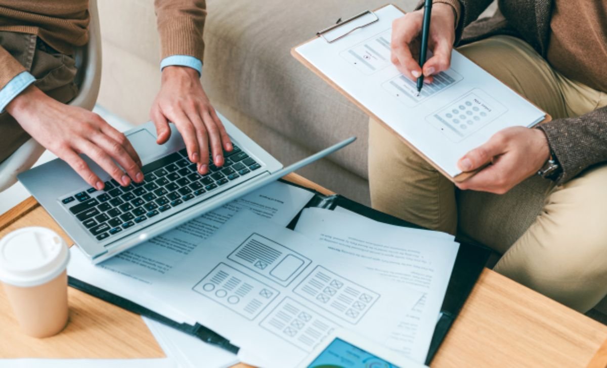 Software design specification defination concept showing professionals reviewing system wireframes and technical documents on laptop and clipboard during planning discussion.